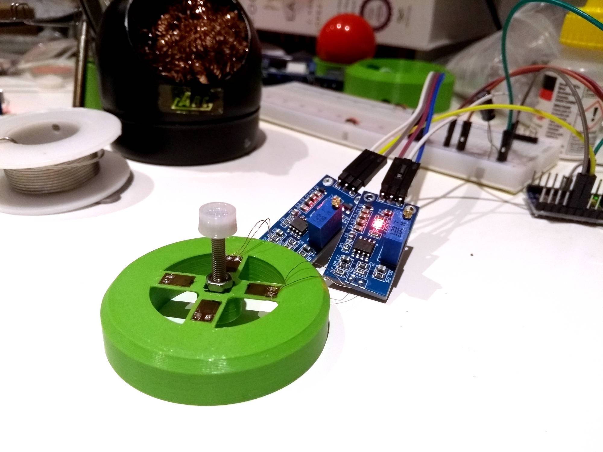 Close-up of a pointing stick prototype. It's got an outer ring for support, with a plus shape in the middle and a screw sticking up out of the centre. There are four strain gauges glued to the plus shape, with two of them wired to two amplifier PCBs with extremely tiny wires.